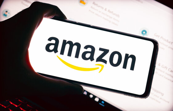 Amazon.  A logo da Amazon na tela de um celular que est&aacute; na m&atilde;o de um homem, e ao fundo  de cena um notebook .Fotografia em ambiente noturno. Site, Com&eacute;rcio eletr&ocirc;nico, online, Venda, Varejo, produto.