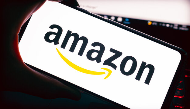 Amazon.  A logo da Amazon na tela de um celular que est&aacute; na m&atilde;o de um homem, e ao fundo  de cena um notebook .Fotografia em ambiente noturno. Site, Com&eacute;rcio eletr&ocirc;nico, online, Venda, Varejo, produto.