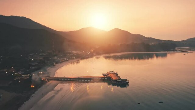 Aerial slow flight over evening beach. Pier in sea, light houses around mountain with trees, slow motion. Budget holiday on Mayreau island in Grenadines, golden hour. Comfortable water taxi backlit.