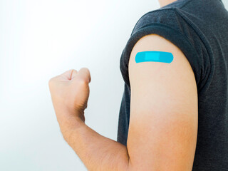 Vaccinations, bandage on vaccinated people concept. Blue bandage on man shoulder who wearing grey shirt and showing strong gesture with fist and arm after vaccination treatment on white background. - obrazy, fototapety, plakaty
