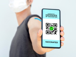 International certificate of vaccination, smart digital passport with QR code on smartphone screen showing by the vaccinated man who wearing medical face mask and blue bandage plaster on his shoulder.