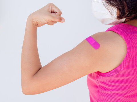 Vaccinations, bandage on vaccinated people concept. Pink bandage on female shoulder who wearing pink sleeveless and face mask and showing strong gesture with fist and arm after vaccination treatment.