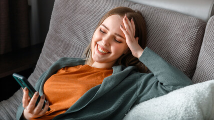 Happy smiling teenage girl laughing texting on phone surfing Internet or chatting using smartphone lying on couch. Young woman use smartphone for leisure enjoyment laughs. Long web banner