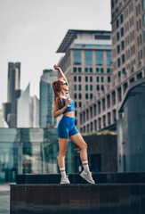 Young slim, flexible, athletic red-haired woman in sportswear, runner stands on city street holding bottle of water and the other hand and looks aside.