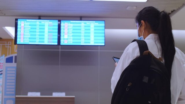 4K Asian tourist women wear protective fack mask with travel bag backpack checking timetable or schedule of flights in Airport COVID-19 Coronavirus pandemic. safe traveling and tourism