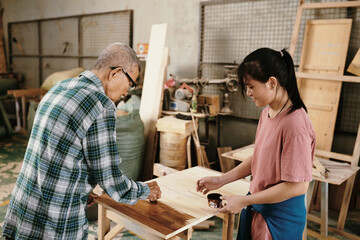 Smiling teenage girl looking at her grandfather covering surface of wooden board with special oil