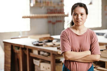 Portrait of young female carpenter crossing arms and looking at camera when standing in workshop