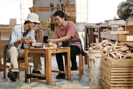Joyful young carpenters enjoying coffee break in workshop, they are discussing picture of creative woodworking project on smartphones