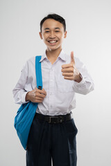 a smiling boy wearing a school uniform and carrying a backpack with thumbs up in isolated background