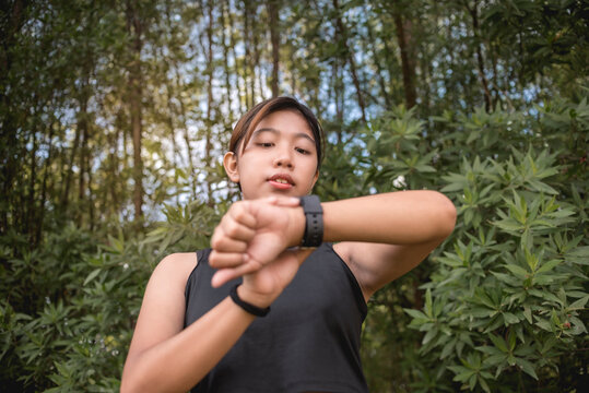 A young asian woman programs or checks the stats on her smartwatch after a trail run through a park or forest.