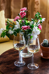 Fototapeta na wymiar Two glasses of white wine and vase with flowers on a wooden table in a cozy room romantic mood