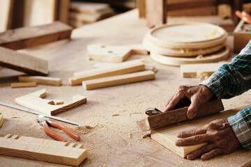 Hands of senior carpenter polishing plank with wooden block at his workbench, selective focus