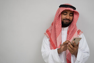 an arabian youth in a turban using a mobile phone wiping the screen while smiling on a plain...