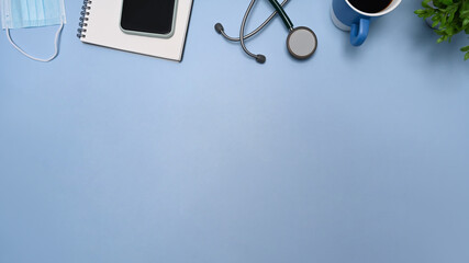 Top view doctor workspace with smart phone, stethoscope, notebook and coffee cup on blue background.