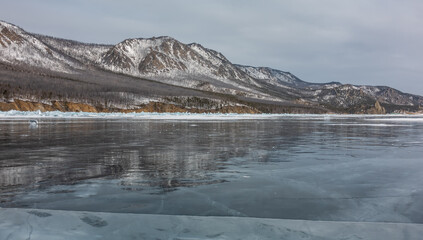 Shards of ice lie on the frozen lake. Blocks of hummocks near the shore. Cracks on smooth ice. Mountain ridge against the sky. Reflection on ice. Baikal