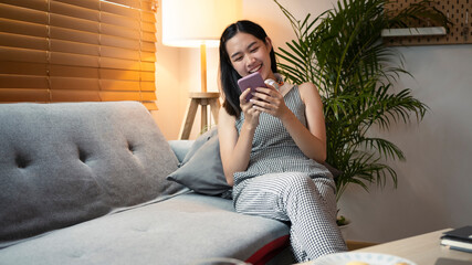 Obraz premium Smiling female sitting on sofa at home and using mobile phone.