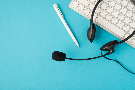 Top view photo of black headphones with microphone pen and white keyboard on isolated pastel blue background with copyspace