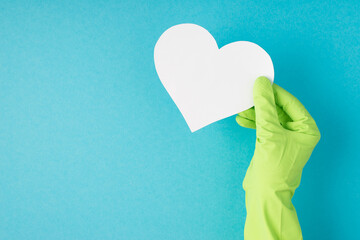 Top view photo of hand in green rubber glove holding white paper heart on isolated pastel blue...