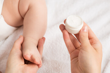 Closeup photo of mother's hands holding newborn's leg and little round cream bottle on isolated white blanket background