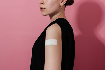 Smiling young woman in t-shirt showing plaster on shoulder after coronavirus vaccine, pink...