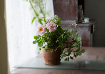 A bouquet of pink roses, geraniums and wildflowers in a clay pot stands on a glass stand on a table in an old house.