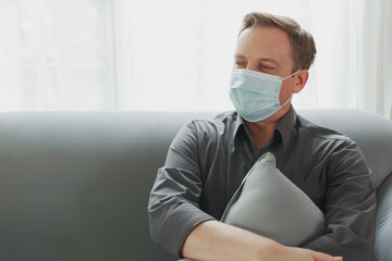 Young attractive Caucasian men wearing protective face mask relaxing on a couch in the afternoon. Happy employee man holding the grey pillow resting with on the sofa during state quarantine