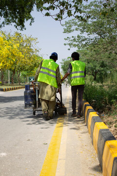 Two blue-collar workers wearing safety jackets are operating a paint-machine to paint / draw road markings.
