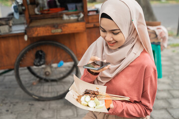 portrait of muslim sate ayam or chicken satay buyer taking photo of her food using smart phone