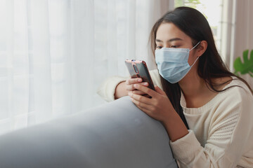 Young Asian woman wearing a face mask stays quarantine at her apartment. Lonely woman using smart phone chatting with her family during social distancing prevent from Covid-19 pandemic crisis.