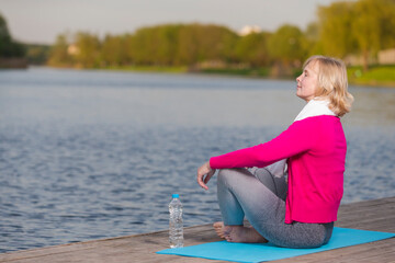 Mature Caucasian Blond Woman During Yoga Practice on Blue Mat At Water Shore Outdoor With Bottle of Water Nearby.