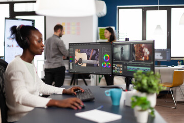 African videographer wearing headset editing movie using post production software working in creative start up agency office. Black editor processing video footage on computer with dual displays