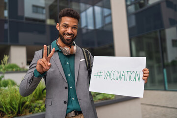 Cheerful young man gesturing V sign with fingers outdoors in city, covid-19 vaccination campaign...