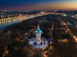 Budapest, Hungary - Aerial view of the illumimnated Margaret Island Open-Air Stage (Margitszigeti Szabadteri Szinpad) water tower at dusk with clear  golden sky and downtown of Budapest at background