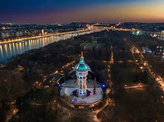 Foto auf Acrylglas Budapest, Hungary - Aerial view of the illumimnated Margaret Island Open-Air Stage (Margitszigeti Szabadteri Szinpad) water tower at dusk with clear  golden sky and downtown of Budapest at background © zgphotography