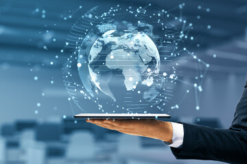 Businessman hand holding tablet with glowing globe interface on blurry office background. Global futuristic innovation concept. Double exposure.
