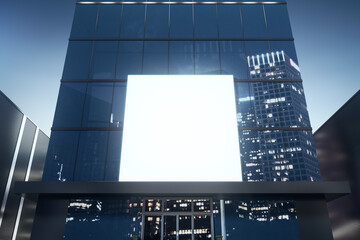 Square white billboard on shiny glass building exterior with reflections at night time. Mock up, 3D...