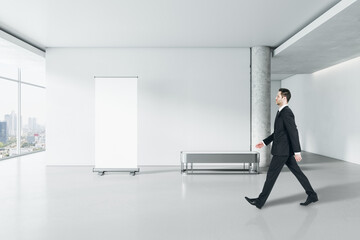 Businessman walking in modern gallery interior with empty poster on concrete wall and window with city view. Mock up.