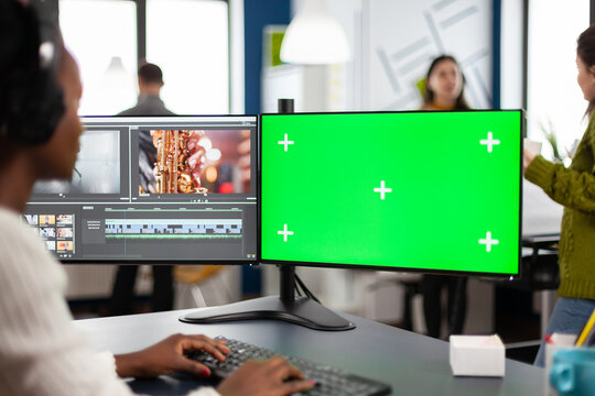 African video editor with headset editing footage using pc with greenscreen, chroma key isolated display sitting in creative agency. Videographer in digital multimedia company processing movie