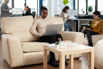 Concentrated business woman reading news on laptop, sitting on couch in busy start up company office, while diverse team analyzes statistics datas. Multiethnic team talking about project.