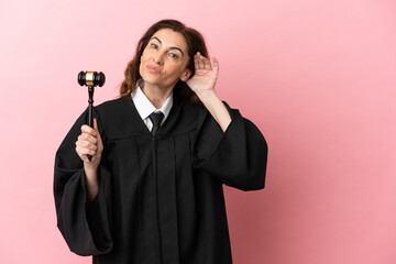 Middle aged judge woman isolated on pink background listening to something by putting hand on the...