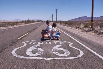 Poster route 66 trip usa. couple posing crouching looking back next to the route 66 sign painted on the road, tourists, travel through usa © jordi