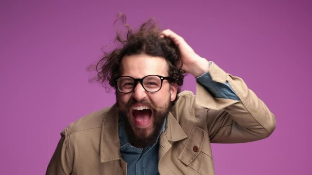 casual unshaved guy in beige jacket with glasses and long beard messing up hair, having fun and laughing while crossing arms on pink background in studio