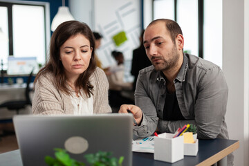 Business man looking at woman colleague and talking about business plan using laptop sitting at table in creative cozy office concentrated on job, while diverse team working in background.