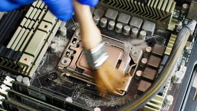 Electronics repair technician in a blue glove sweeps dust off the computer processor on the motherboard of a desktop computer from old coolant with a special brush. Close up