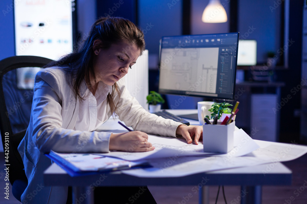 Poster overworked woman architect checking and matching blueprints sitting at office desk in front of compu