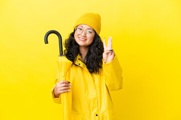 Asian woman with rainproof coat and umbrella isolated on yellow background happy and counting three with fingers