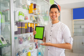 smiling man holding a tablet with hand gesture pointing at tablet screen near glass display case on smartphone shop background