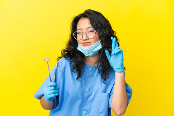 Young asian dentist holding tools over isolated background with fingers crossing and wishing the best