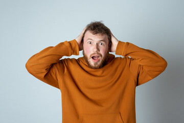 Image of shocked bearded modern young man in stylish hoodie, holding hands on head and feeling distressed