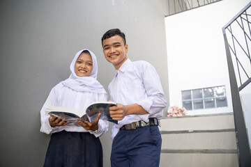 two teenagers in junior high school uniforms study together using a book while standing on the stairs in the room